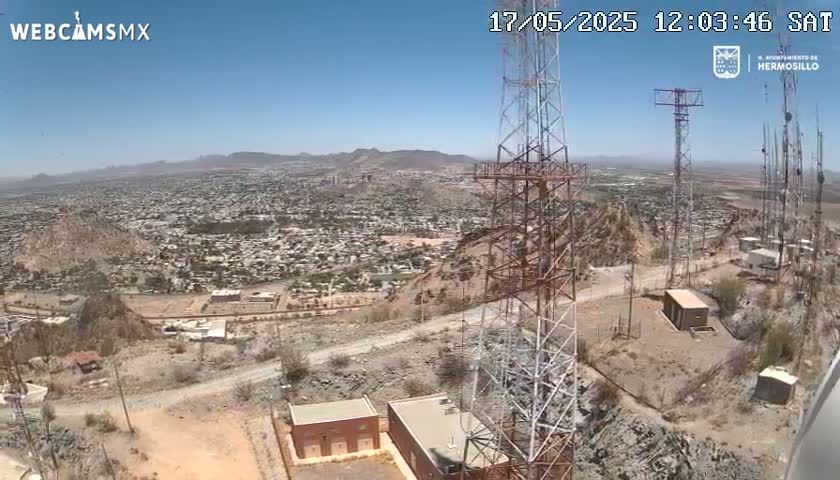A high-angle, long shot on a clear, sunny day shows a sprawling city nestled in a mountainous region, with several tall communication towers and smaller buildings visible in the foreground.