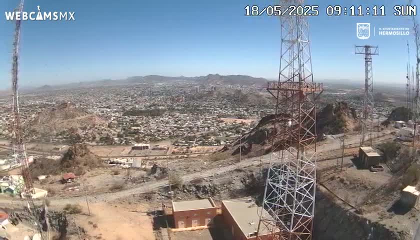 A high-angle view on a clear, sunny day shows a sprawling city in a valley below, with several tall communication towers situated on a rocky hillside overlooking the cityscape.