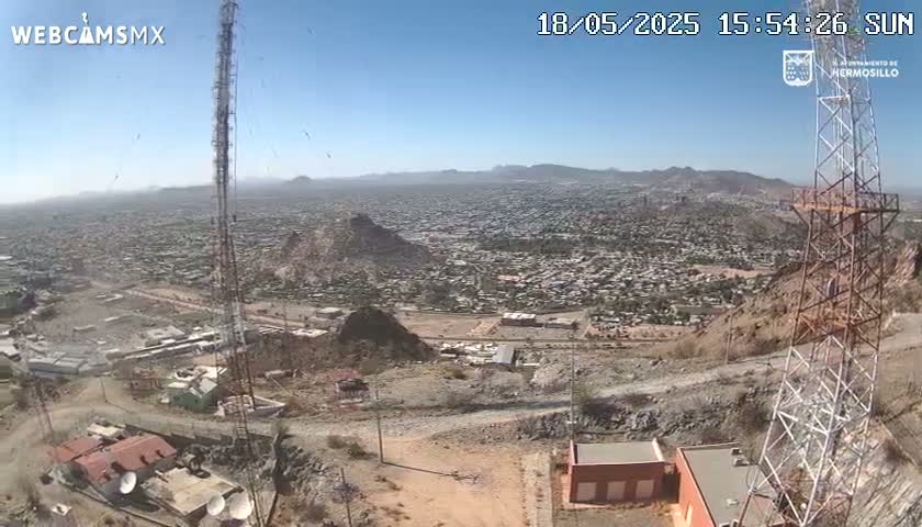 A high-angle, long shot on a sunny day shows a sprawling city extending to distant mountains, viewed from a rocky hillside with communication towers and buildings nearby.