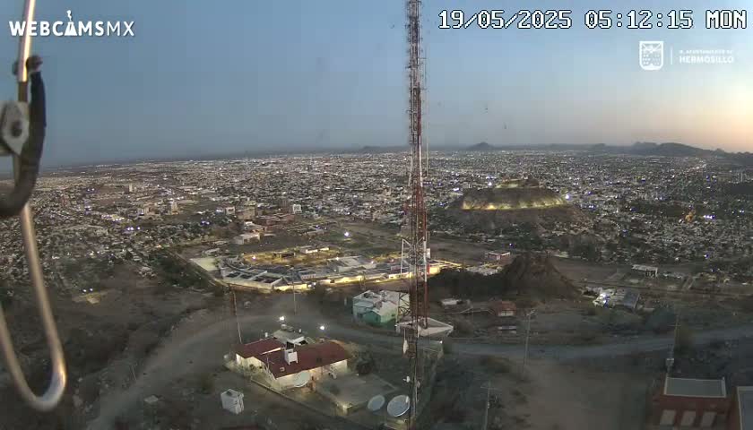 An aerial twilight view of a city at night, showing a large communication tower near a building complex, with hills and mountains in the background.