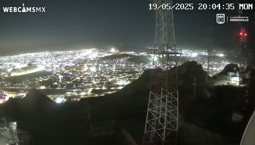 A nighttime, high-angle view shows a sprawling city illuminated below, as seen from a hilltop with two tall communication towers in the foreground.