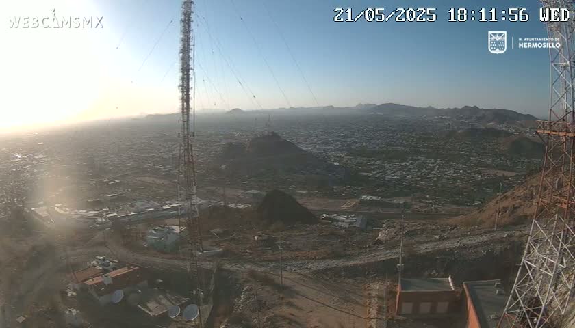 A hazy sunrise illuminates a sprawling city viewed from a high vantage point, with communication towers and surrounding terrain visible in the foreground.