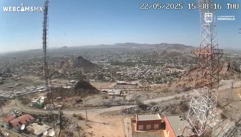 A high-angle, long shot on a clear, sunny day shows a sprawling city nestled in a mountainous desert landscape, with two tall communication towers prominently featured in the foreground.