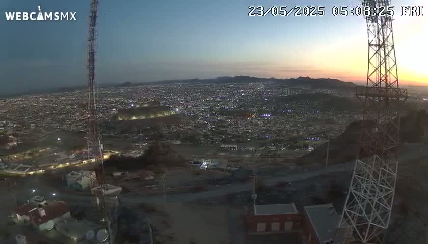 An aerial view at sunset shows a sprawling city illuminated at night, with a tall communication tower in the foreground on a hill.