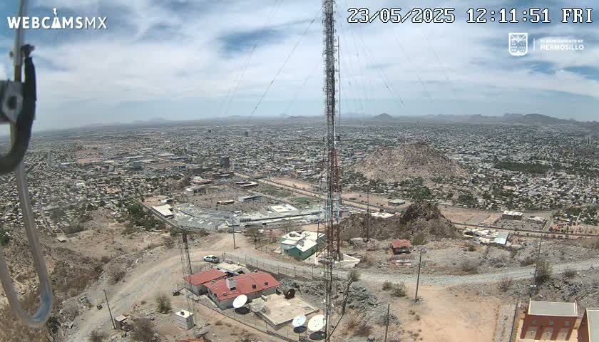 A mostly clear day reveals a sprawling city viewed from a hilltop, featuring a tall communication tower and several buildings near the camera's vantage point.