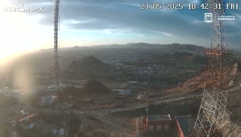 A high-angle, long shot of a city at sunset, viewed from a hilltop with two tall communication towers and various equipment, under mostly clear skies.
