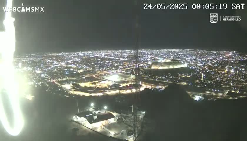 A nighttime, high-angle view shows a sprawling city illuminated with lights, partially obscured by hills, and a tall tower in the foreground.