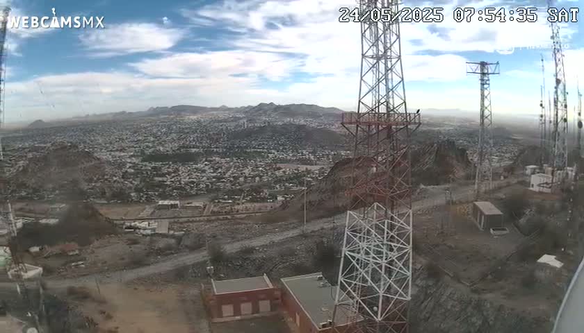 A high-angle, long shot on a partly cloudy day shows a sprawling city below a hilltop with several communication towers and buildings.