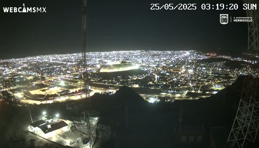 A nighttime, high-angle view shows a sprawling city illuminated by countless lights, situated below a dark hilltop.