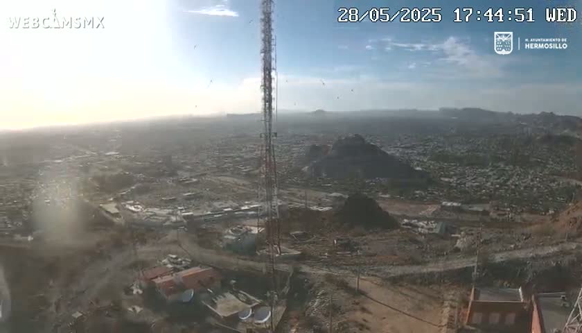 A mostly sunny, high-angle view shows a sprawling city below a tall communication tower on a dusty, hilly landscape.