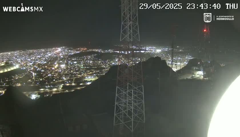 A nighttime view from a hilltop shows a sprawling city illuminated below, with communication towers prominent in the foreground under a clear sky.