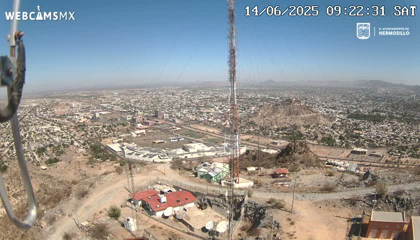 A high-angle, long shot on a clear, sunny day shows a sprawling city below a hilltop with several buildings, communication towers, and satellite dishes; a rocky, desert landscape surrounds the structures.