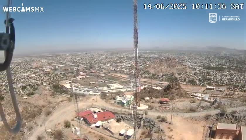 A high-angle, long shot on a clear, sunny day shows a sprawling city below a hilltop with several buildings, communication towers, and satellite dishes; a rocky, desert landscape surrounds the structures.