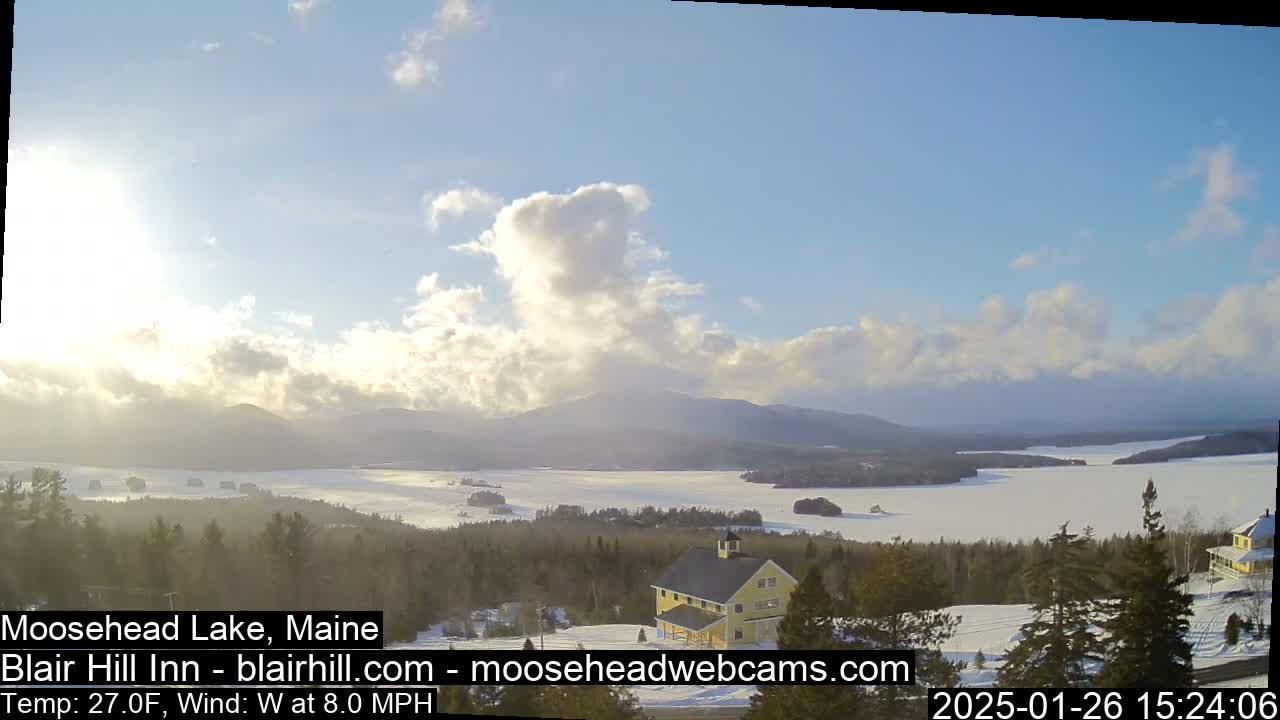 A partly cloudy, wintery scene overlooks a snow-covered lake and distant mountains, with a yellow house situated in the foreground amongst evergreen trees.