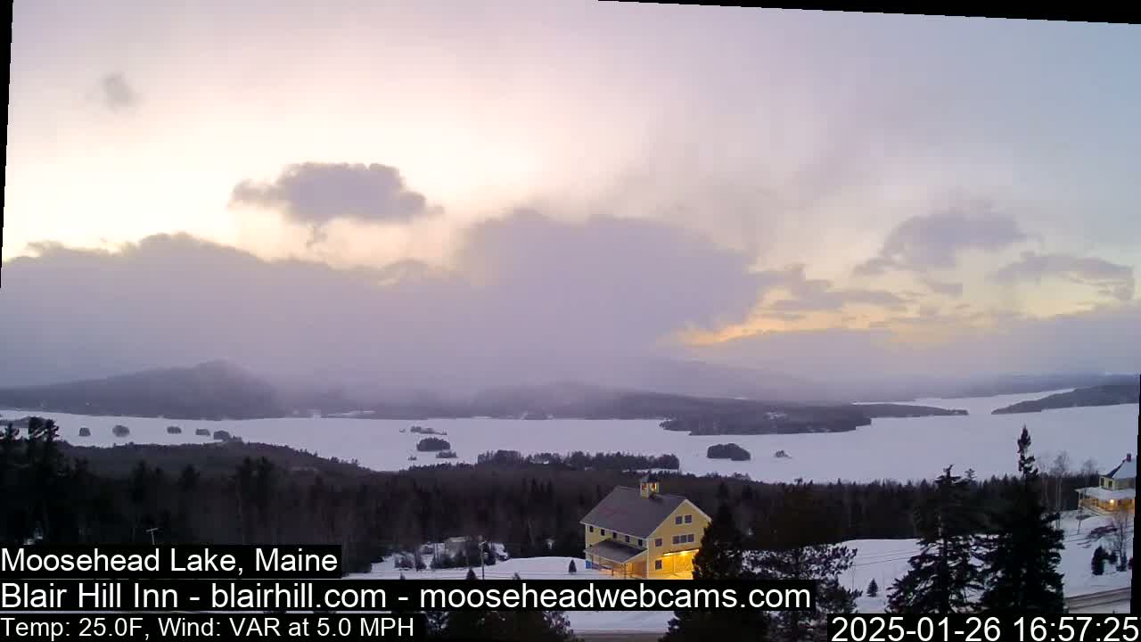 A partially frozen lake is visible from a snowy hilltop at dusk under a cloudy sky.