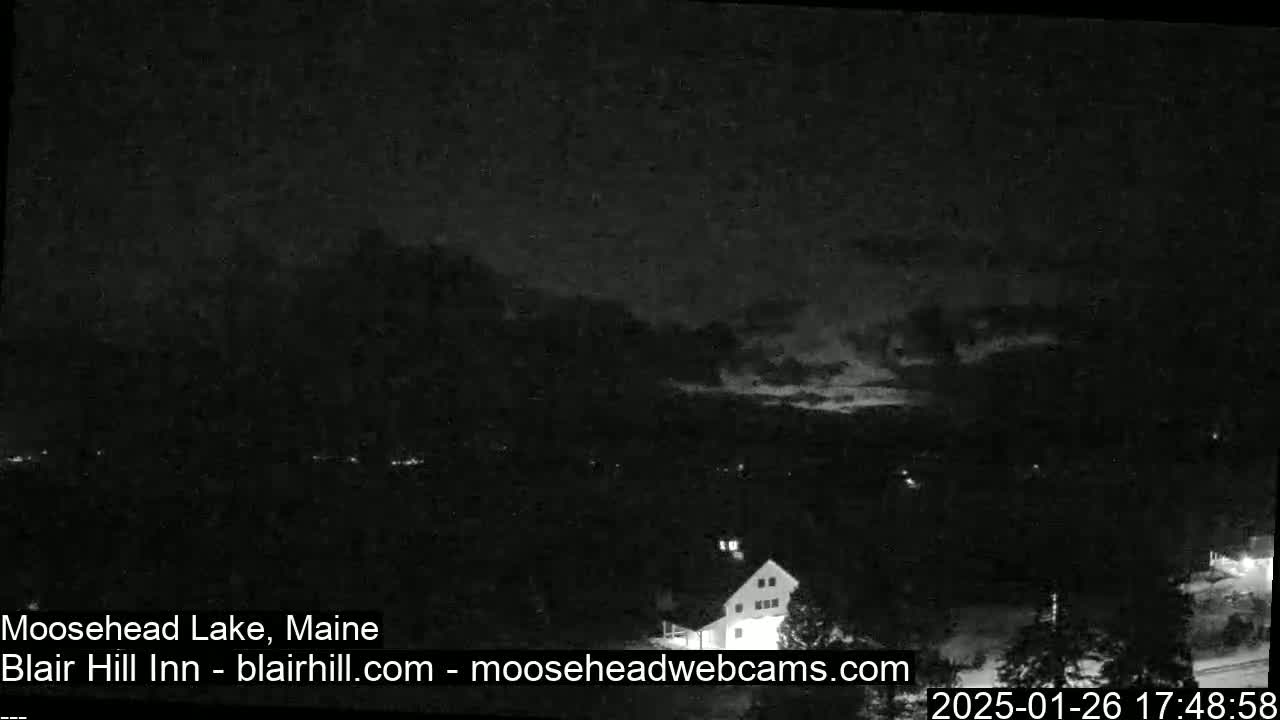 A nighttime, low-light image shows a dark landscape with a building visible in the foreground under a mostly cloudy sky.