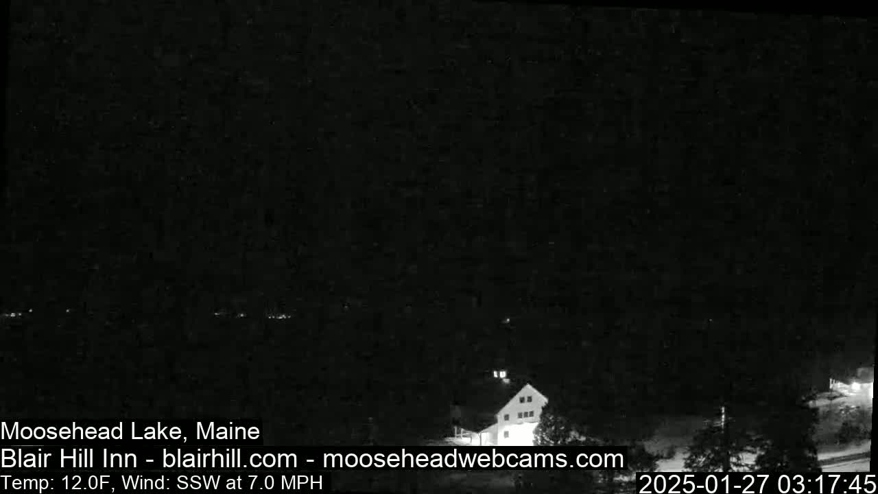 A nighttime, low-light image shows a dark landscape with a building and some vegetation, under a clear, dark sky with a south-southwesterly wind at 7.0 mph and a temperature of 12.0°F.