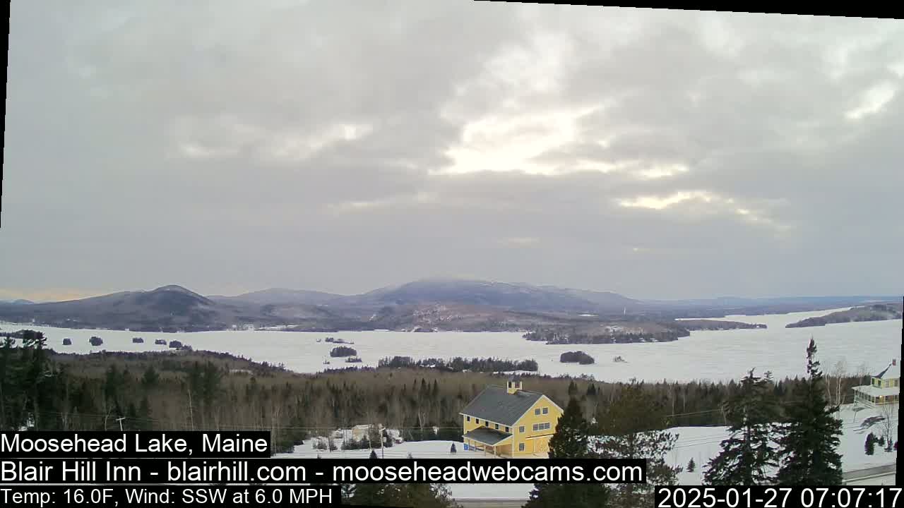 A mostly cloudy, cold day overlooks a partially frozen lake and distant mountains with a yellow building in the foreground.