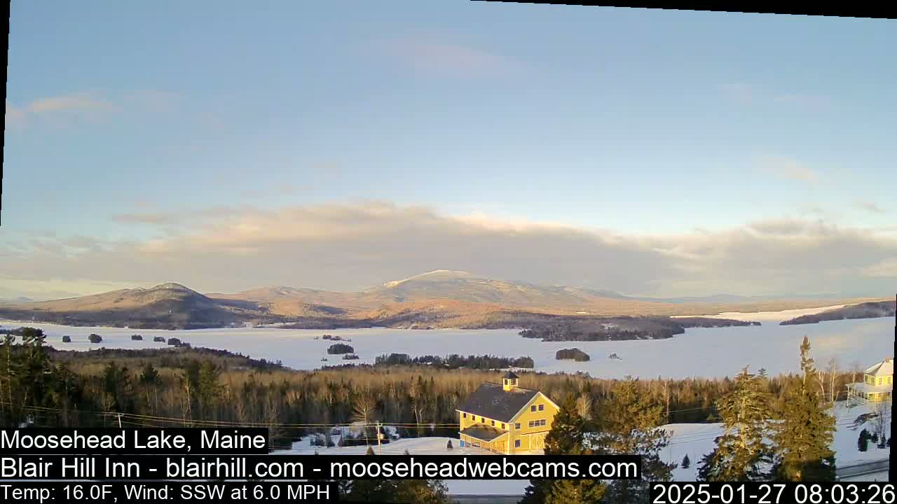 A partly cloudy, cold day with a light south-southwesterly wind overlooks a snow-covered lake and distant mountains, featuring a yellow building in the foreground.