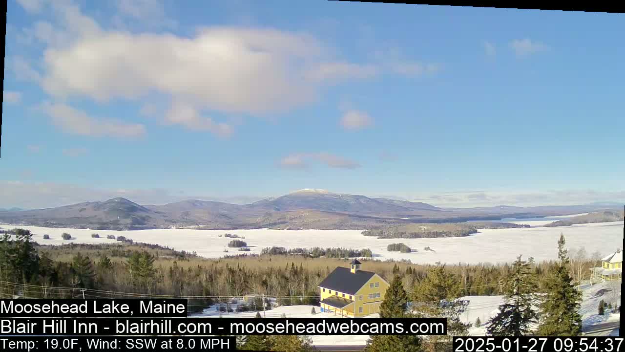 A partly cloudy, cold day shows a frozen lake and distant mountains, viewed from a hilltop with a yellow building in the foreground.