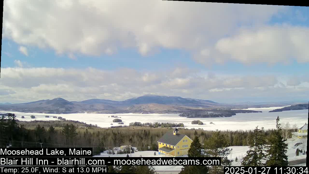 A partly cloudy day shows a partially frozen lake and distant mountains, with a yellow building in the foreground and a snow-covered landscape.