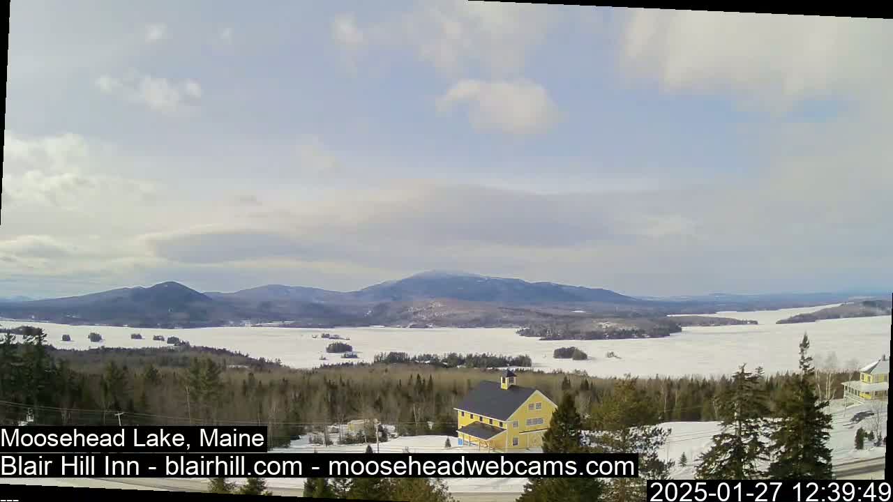 A partly cloudy day reveals a mostly frozen lake and distant mountains, with a yellow building visible in the foreground.