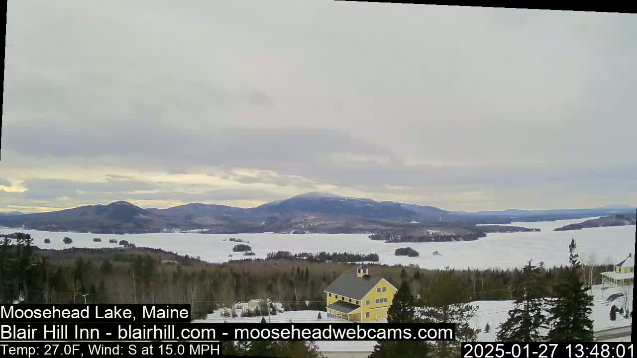A partly frozen lake is visible from a snowy vantage point, with a yellow building and dark, snow-covered evergreen trees in the foreground, under a mostly cloudy sky.
