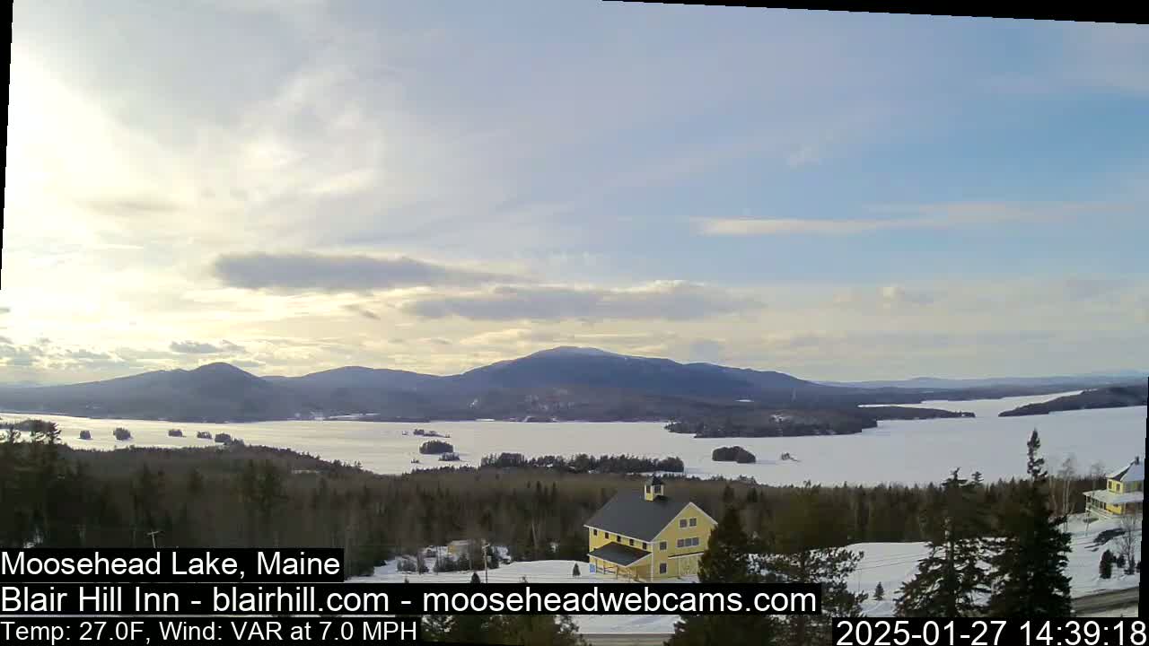 A partly cloudy day reveals a frozen lake nestled between snow-covered hills and mountains, with a yellow building visible in the foreground.