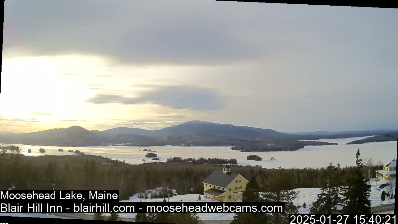 A partly cloudy sunset over a snow-covered lake and forested hills, with a yellow building visible in the foreground.
