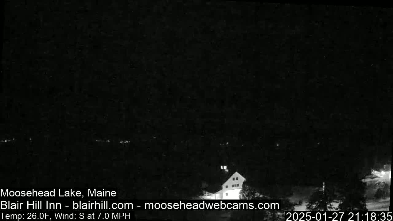 A dark nighttime view of a lake and a building with some faint lights, under a clear, dark sky with a south wind at 7 mph and a temperature of 26 degrees Fahrenheit.