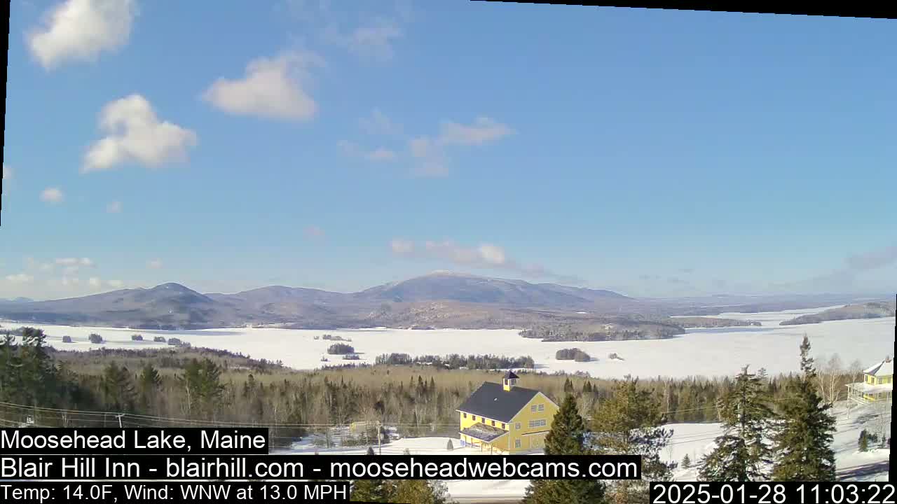 A partially frozen lake is seen from a snowy hillside with a yellow building and distant mountains under a mostly clear, sunny sky.