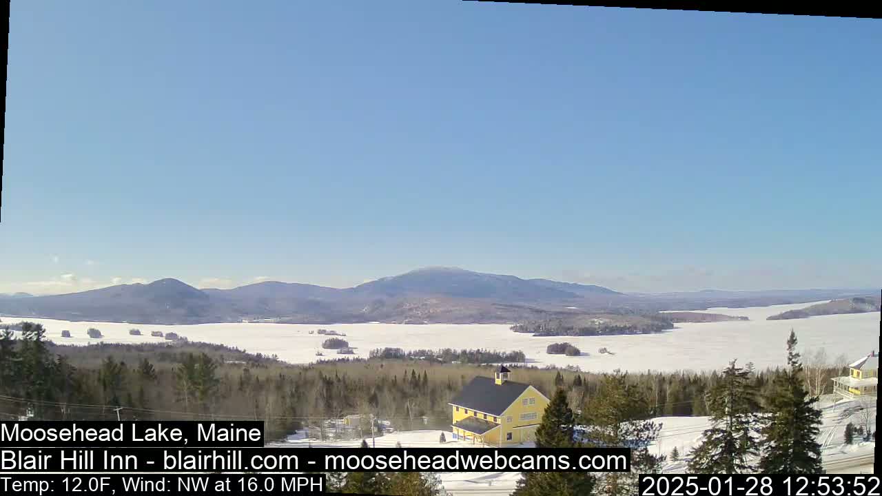 A partly frozen lake stretches to snow-covered mountains under a clear blue sky, with a yellow building and evergreen trees in the foreground, and a northwest wind at 16 mph.