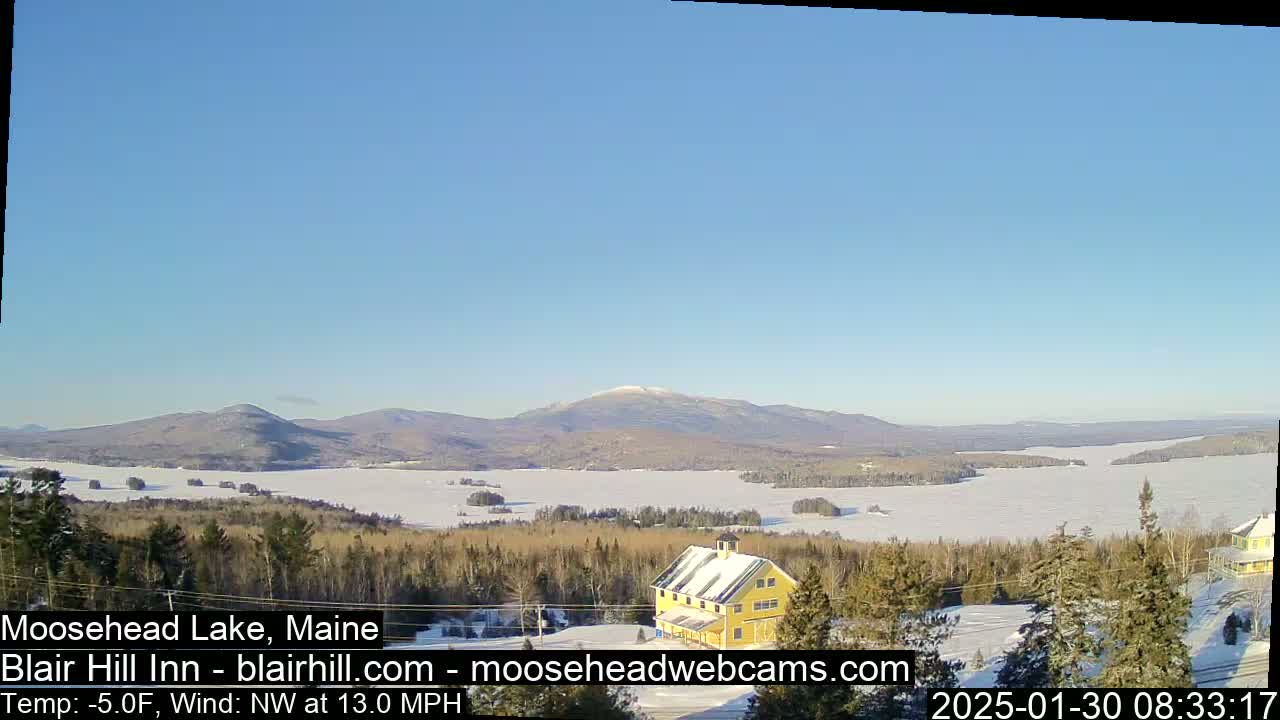 A partially frozen lake is seen from a snowy hilltop, with a yellow building, distant snow-capped mountains, and a clear blue sky under a northwest wind of 13 mph and a temperature of -5°F.