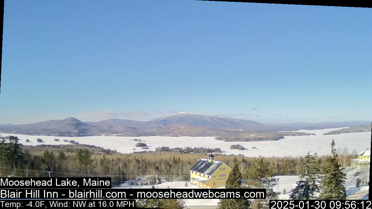 A snow-covered lake and distant mountains are visible under a clear blue sky, with a yellow building in the foreground and a northwest wind of 16 mph at -4°F.