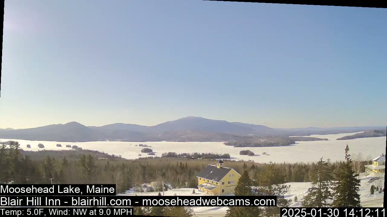 A partially frozen lake is visible from a snowy hilltop, with a yellow building nearby, mountains in the background, and a clear, sunny sky.