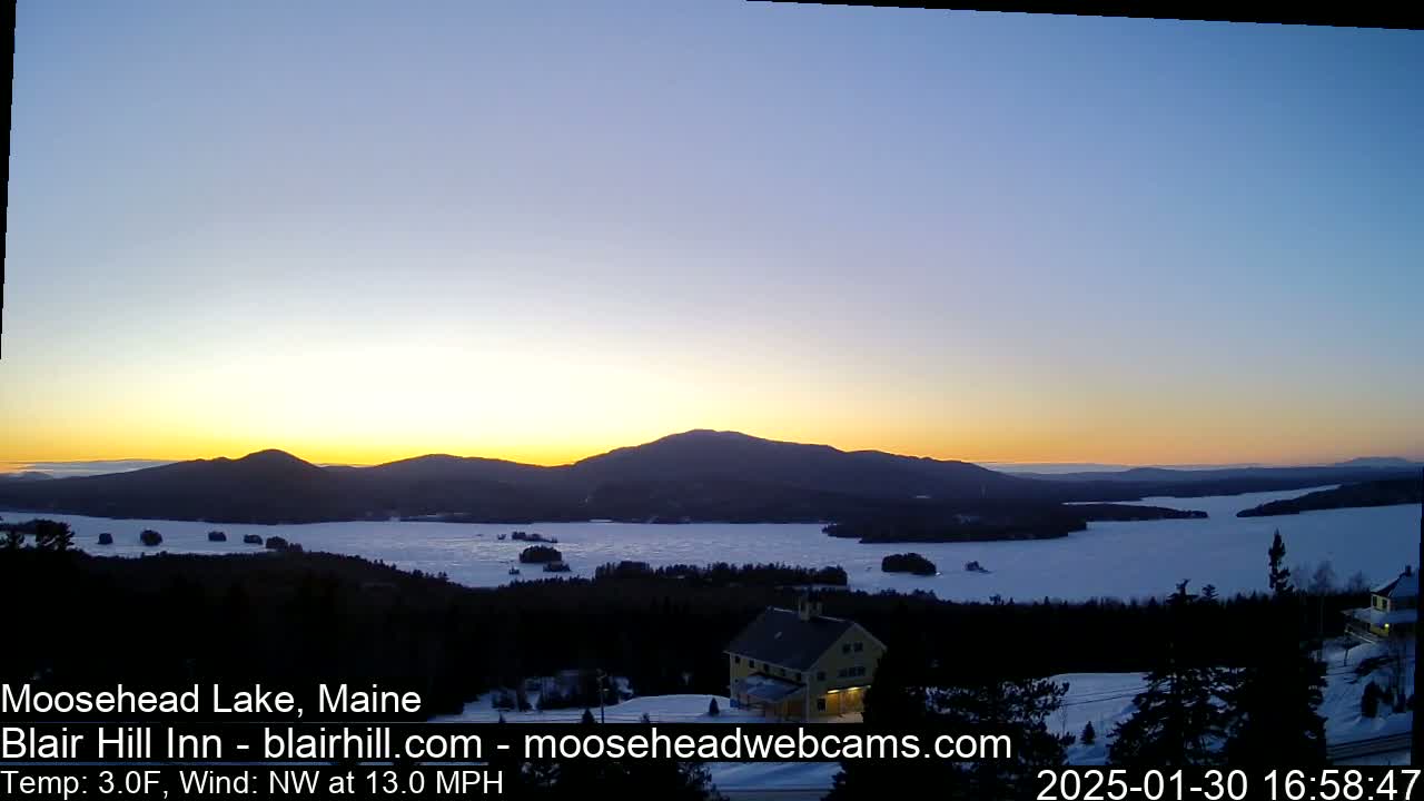 A partially frozen lake at sunset is viewed from a hilltop, with a building visible in the foreground and mountains in the background, under a clear sky and a northwest wind of 13 mph.