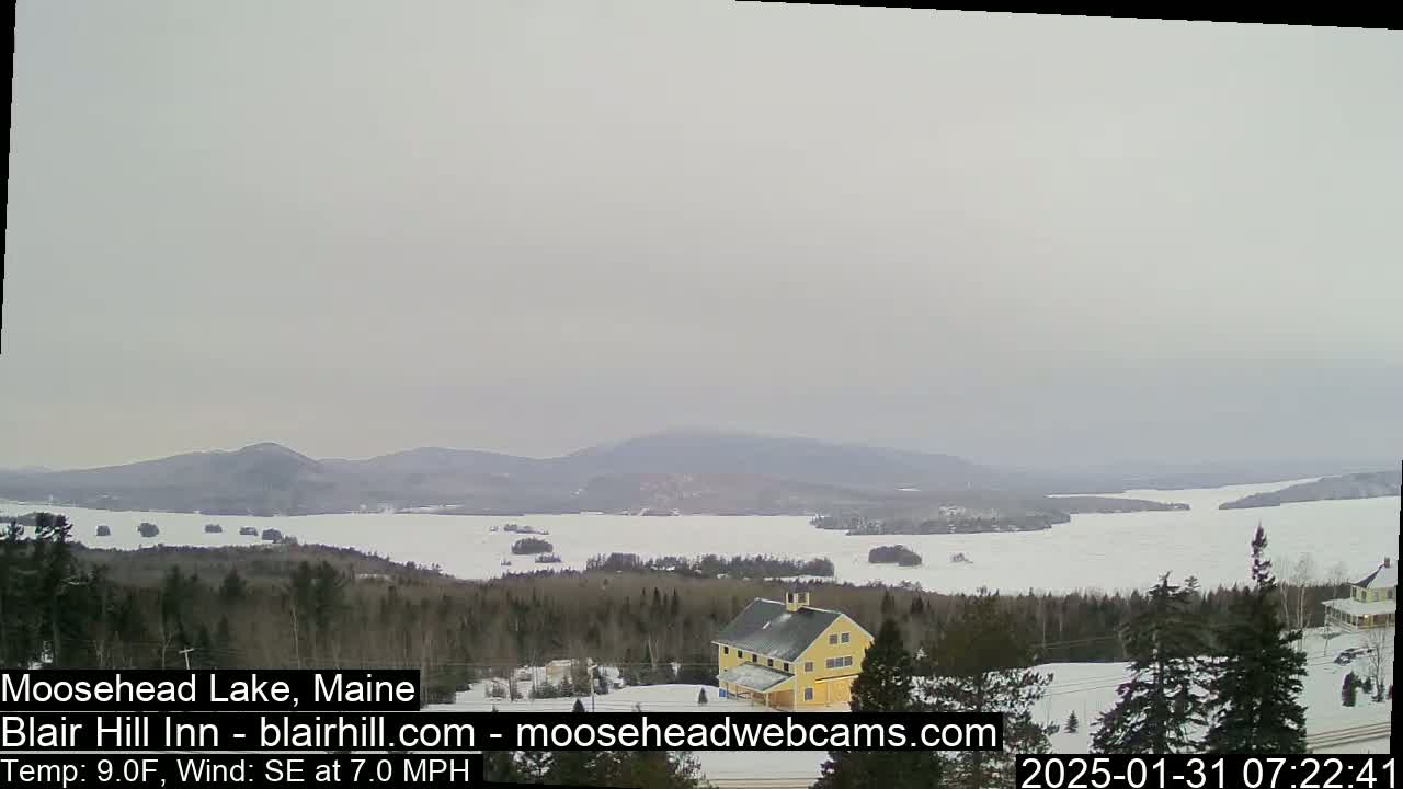 A partially frozen lake is visible from a snowy, wooded hillside, with a yellow building and distant mountains under an overcast sky.