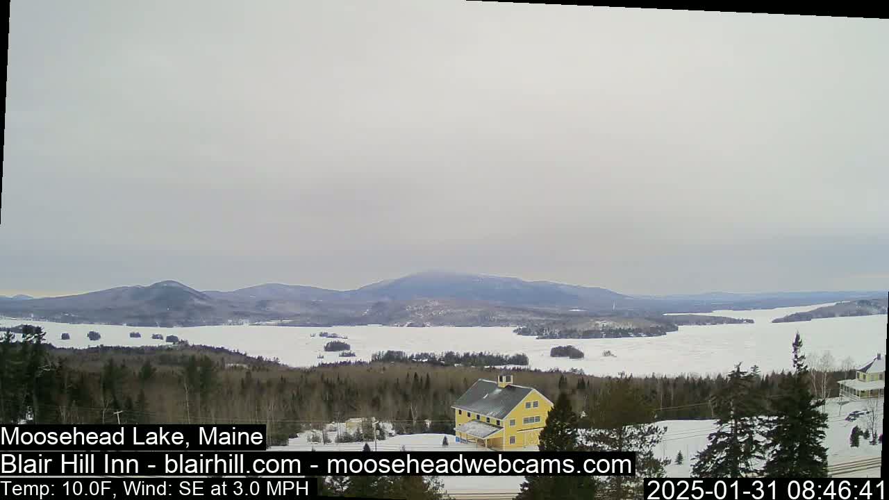 A partly frozen lake stretches out to distant mountains under an overcast sky, with a yellow building visible in the foreground.
