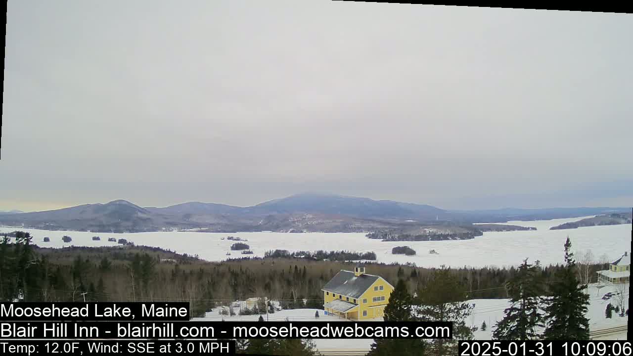 A partly frozen lake is visible from a snowy hillside, with a yellow building and distant mountains under an overcast sky.