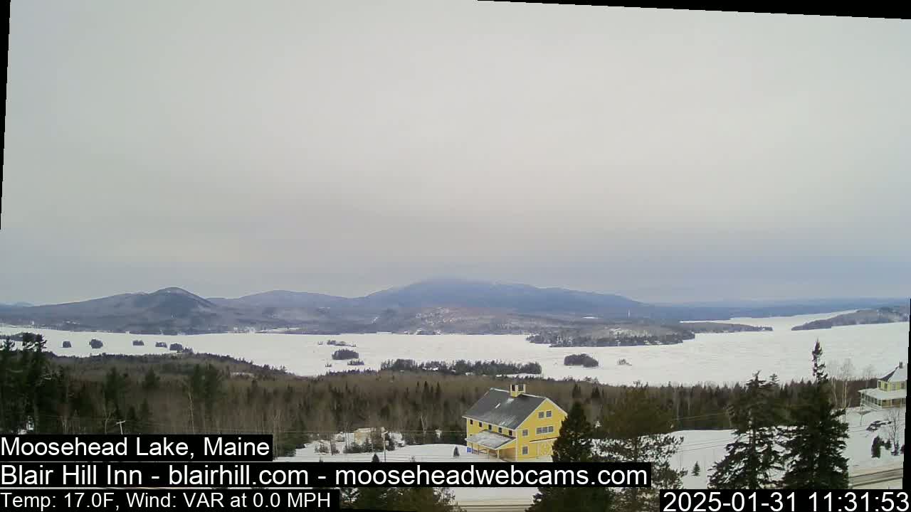 A partially frozen lake stretches to distant mountains under an overcast sky, with a yellow building situated in the foreground among evergreen trees.