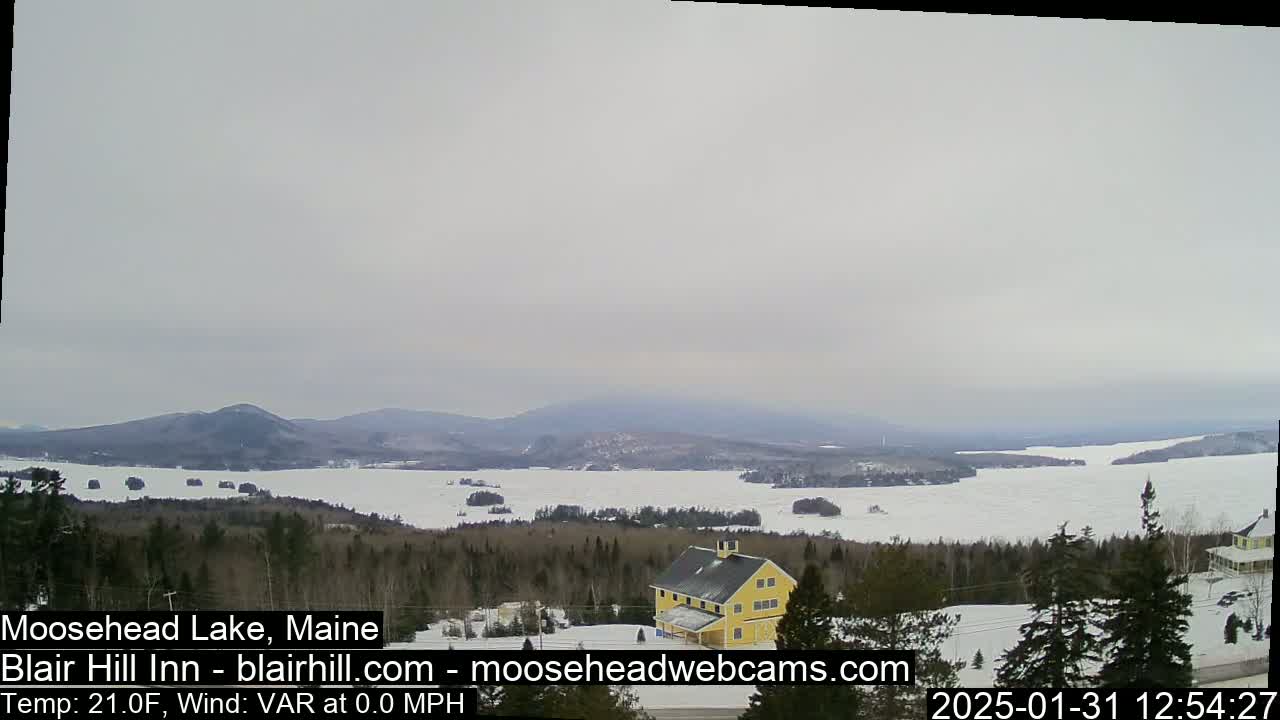A partially frozen lake stretches to distant mountains under an overcast sky, with a yellow building nestled in a snow-covered wooded area in the foreground.