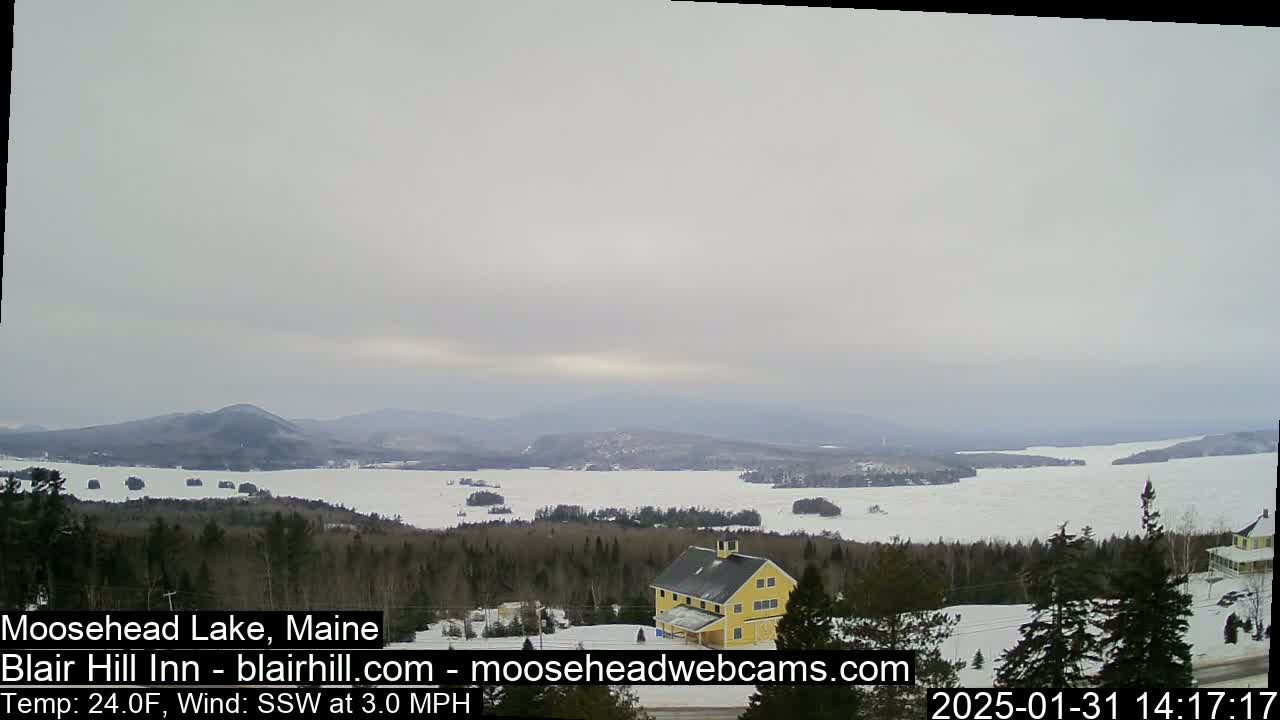 A partially frozen lake is visible from a snowy, wooded hillside, with a yellow building in the foreground under an overcast sky.