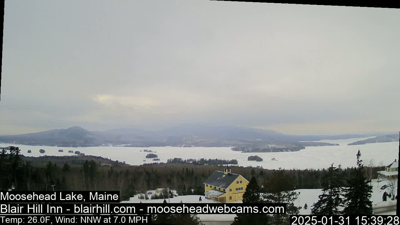 A partially frozen lake stretches to distant mountains under an overcast sky, with a yellow building and snow-covered trees in the foreground.