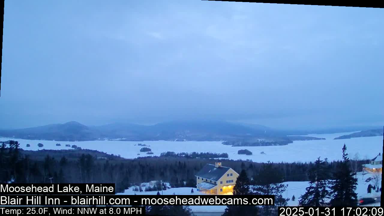 A partially frozen lake is visible from a snowy hilltop, with a building and distant mountains under a pale, cloudy sky.