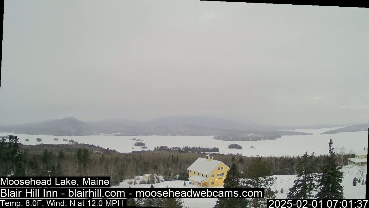 A snow-covered landscape shows a frozen lake with a yellow building and evergreen trees in the foreground under an overcast sky.