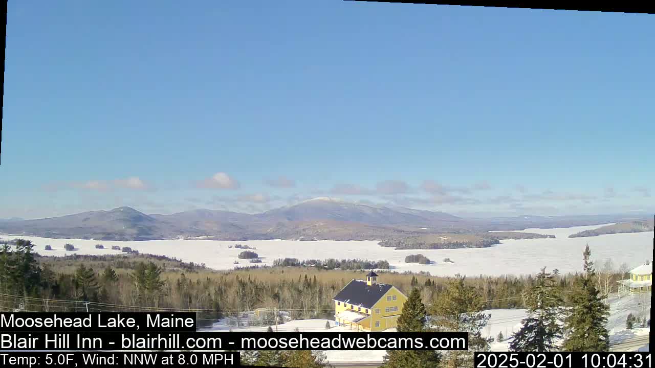 A mostly sunny, cold day shows a partially frozen lake and distant mountains viewed from a snowy hilltop with a yellow building in the foreground.