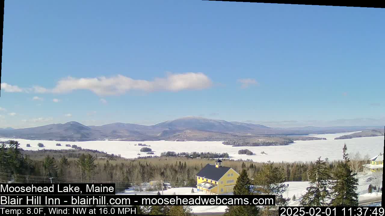 A partly cloudy, cold day shows a frozen lake and distant mountains, with a yellow building nestled in a snowy evergreen forest in the foreground.