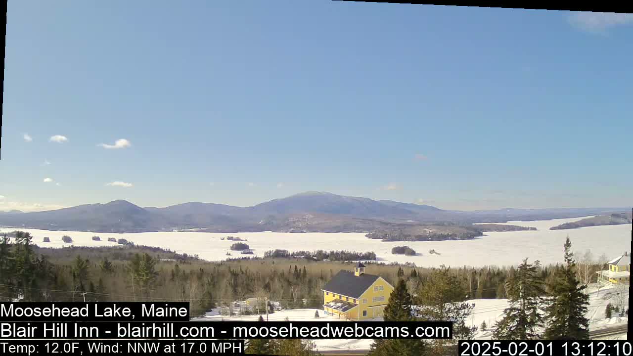 A partly frozen lake is visible under a clear blue sky, with snow-covered land, evergreen trees, and a yellow building in the foreground, and mountains in the background.