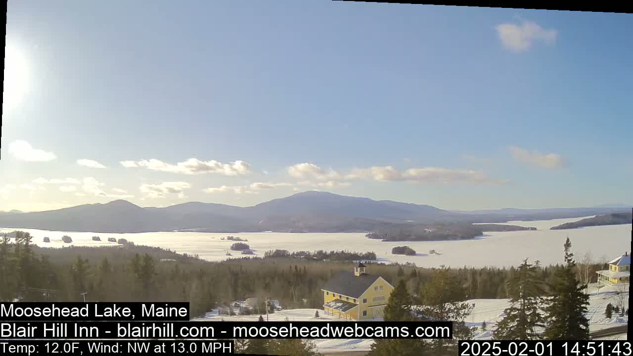 A partly cloudy, cold day reveals a snow-covered lake and distant mountains, with a yellow building nestled in a snow-covered forest in the foreground.