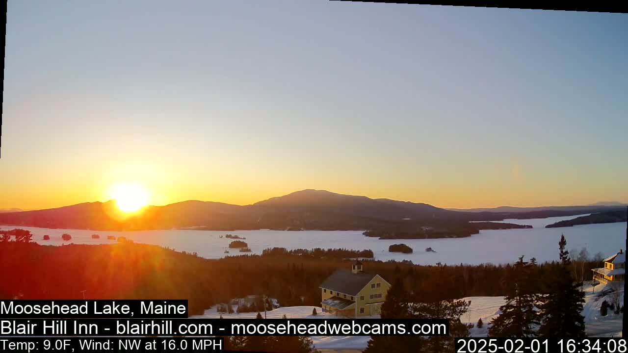 A sunset over a snow-covered lake and mountains, with a yellow building visible in the foreground, under a clear sky and a northwest wind.